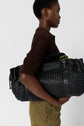 Woman in a brown knit top and olive pants holds a large black woven leather bag with gold-toned hardware against a plain light gray background.