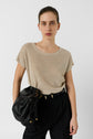 A person with slicked-back hair wears a beige short-sleeve top, black pants, and carries a black leather shoulder bag with gold accents, standing against a plain white background.