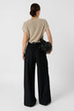 Woman standing with back to camera, wearing a light beige short-sleeve top tucked into high-waisted black wide-leg pants, holding a black bag on her left arm. Her hair is pulled back in a neat bun.