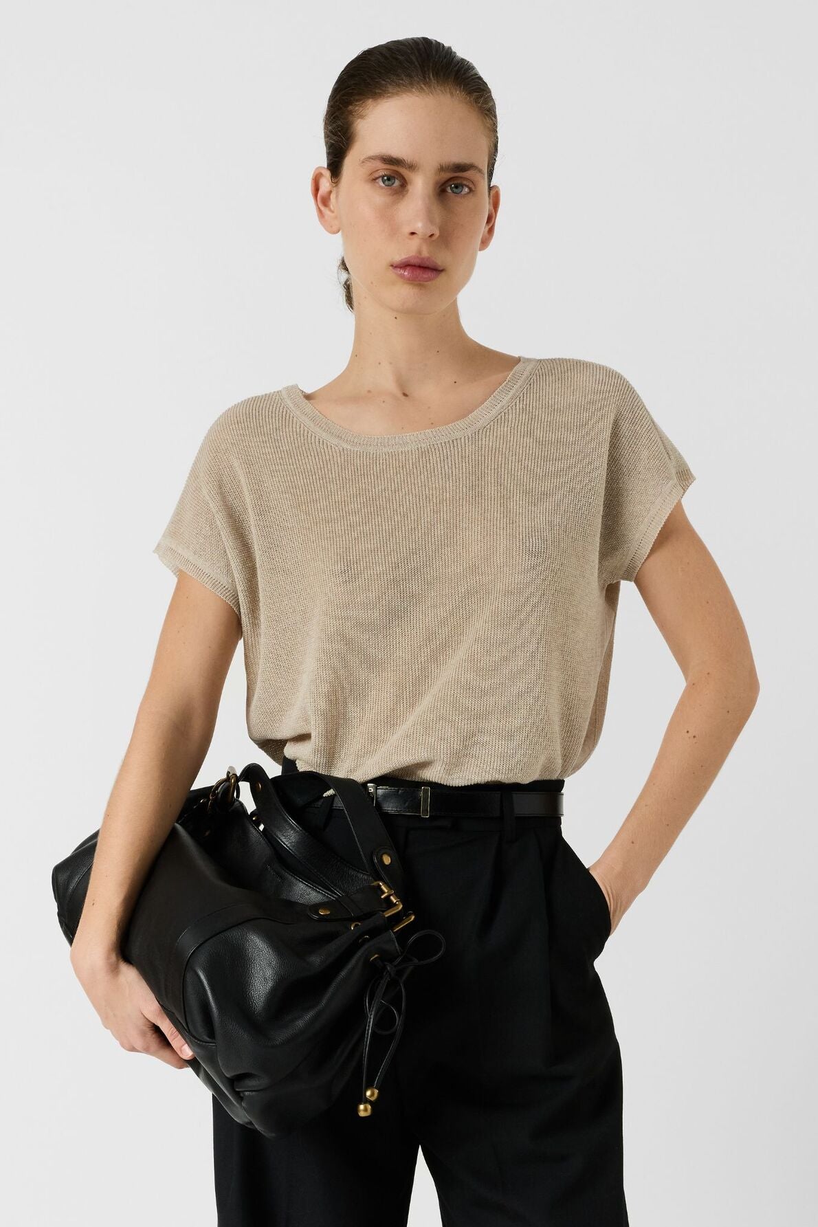 A person with slicked-back hair wears a beige short-sleeve shirt and high-waisted black pants, holding a large black bag on one arm against a plain light background.
