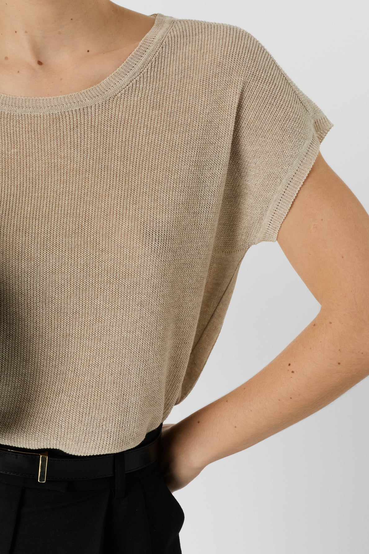 A person wearing a beige short-sleeve knit top tucked into high-waisted black pants, with one hand on their hip, against a plain light background.