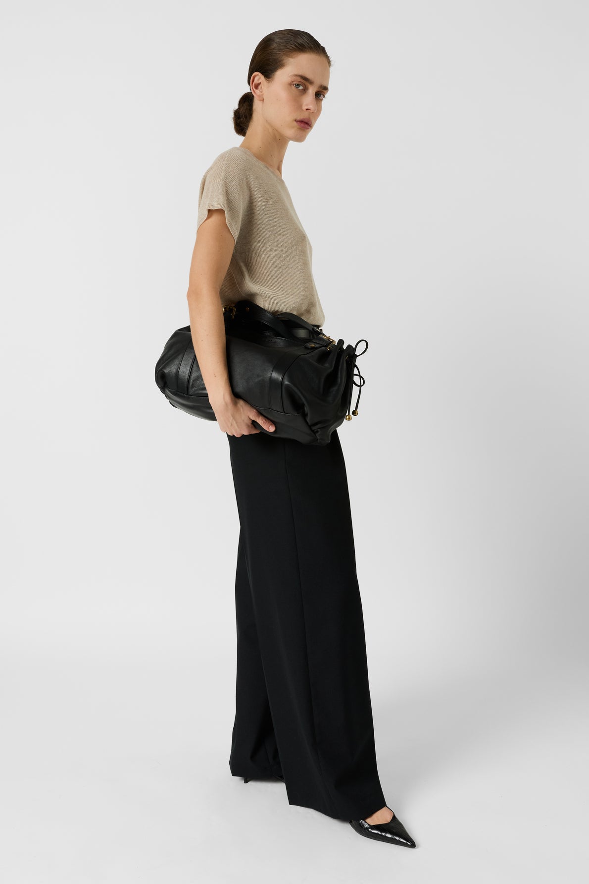 A person wearing a beige short-sleeve top, black wide-leg pants, and black pointed shoes stands against a plain white background, holding a large black bag under one arm.