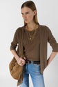A person wearing a brown cardigan, layered gold necklaces, rings, and carrying a brown suede fringe bag stands against a plain background, looking to the side with a confident expression.