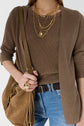 A person wears a brown knit top and cardigan with blue jeans and a dark braided belt. They have a brown suede fringe bag over their shoulder, multiple gold chain necklaces with a heart charm, and a large statement gold ring on their finger.