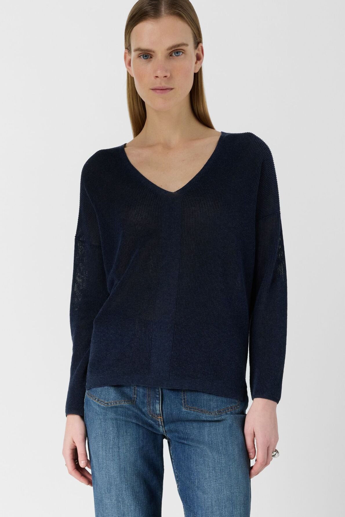 A person with straight, light brown hair wears a dark blue V-neck sweater and blue jeans, standing against a plain white background.