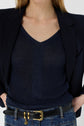 Woman wearing a navy V-neck top, black pinstripe blazer draped over her shoulders, blue jeans, and a black belt with a gold buckle, standing with hands in her pockets.