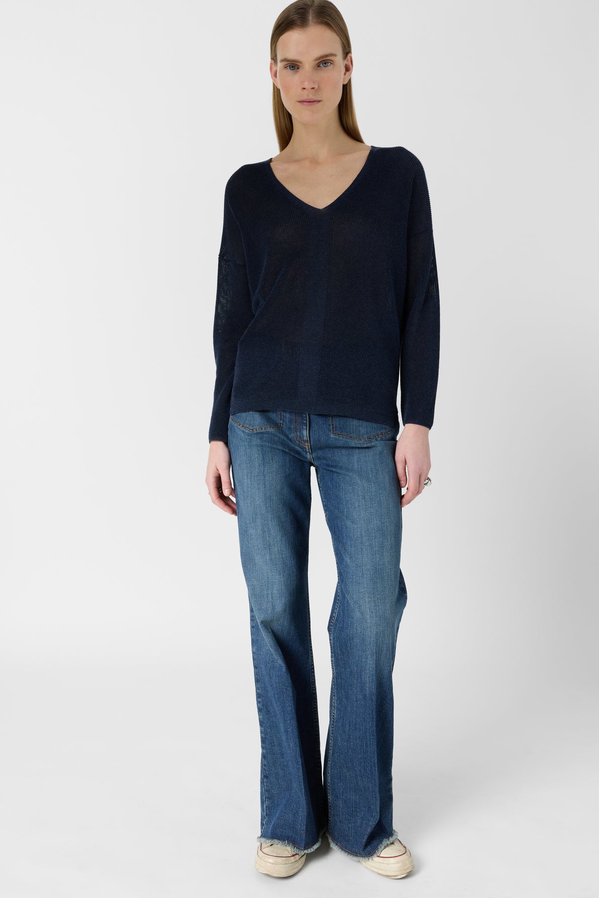 A person stands facing forward, wearing a dark blue V-neck sweater, blue flared jeans, and beige shoes, against a plain white background.