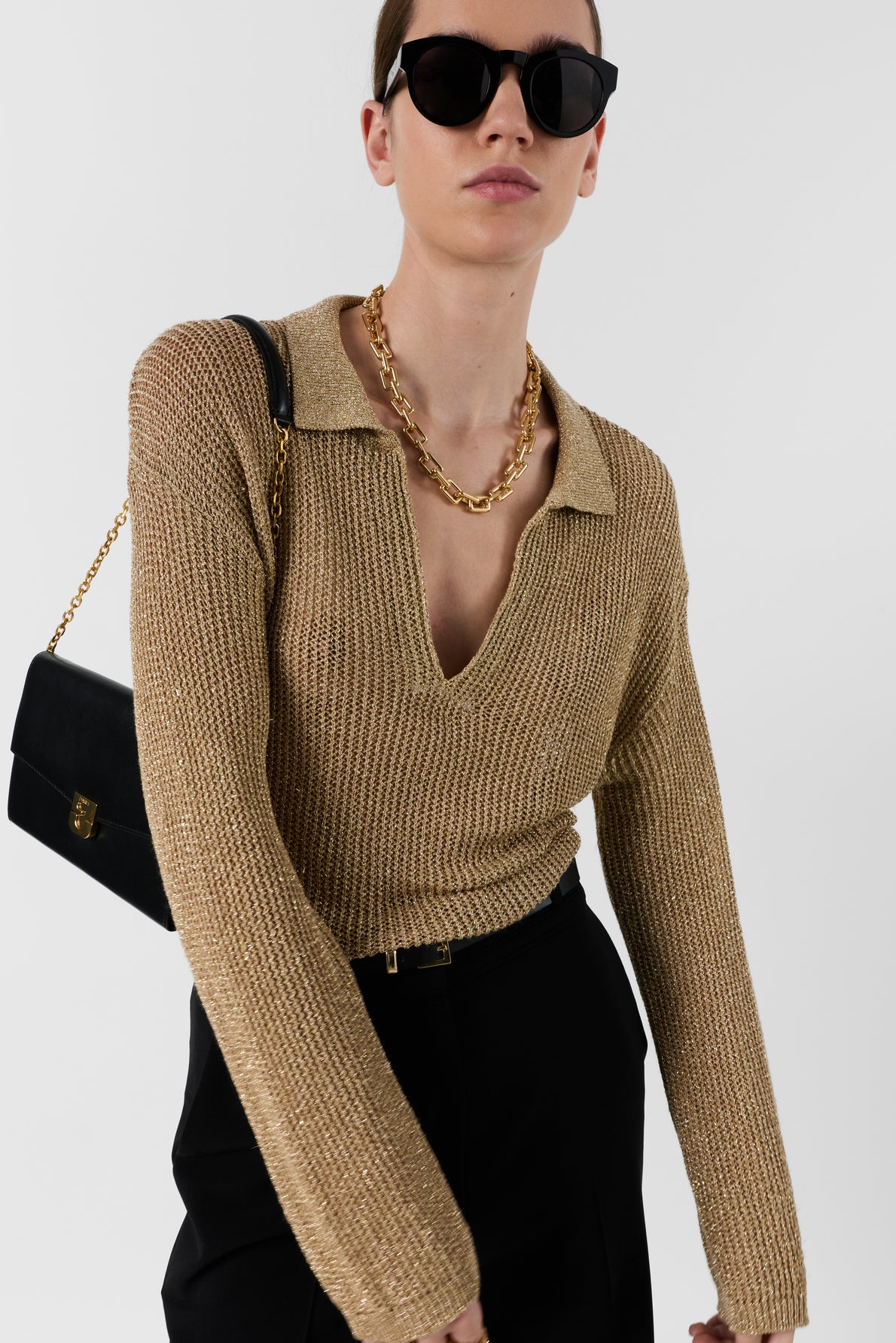 A person wearing sunglasses, a pull Lailah gold knit sweater with a deep collar in maille ajourée, a chunky gold necklace, black pants, and a black shoulder bag with a gold chain, posing against a white background.