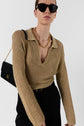 A person wearing sunglasses, a pull Lailah gold knit sweater with a deep collar in maille ajourée, a chunky gold necklace, black pants, and a black shoulder bag with a gold chain, posing against a white background.