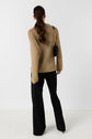 A woman with long brown hair in a ponytail, wearing a tan pull Lailah en maille ajourée, black wide-leg pants, black heeled shoes, and carrying a black shoulder bag, stands facing away from the camera against a white background. The look exudes understated elegance with refined details inspired by pièces fabriqué en Italie.