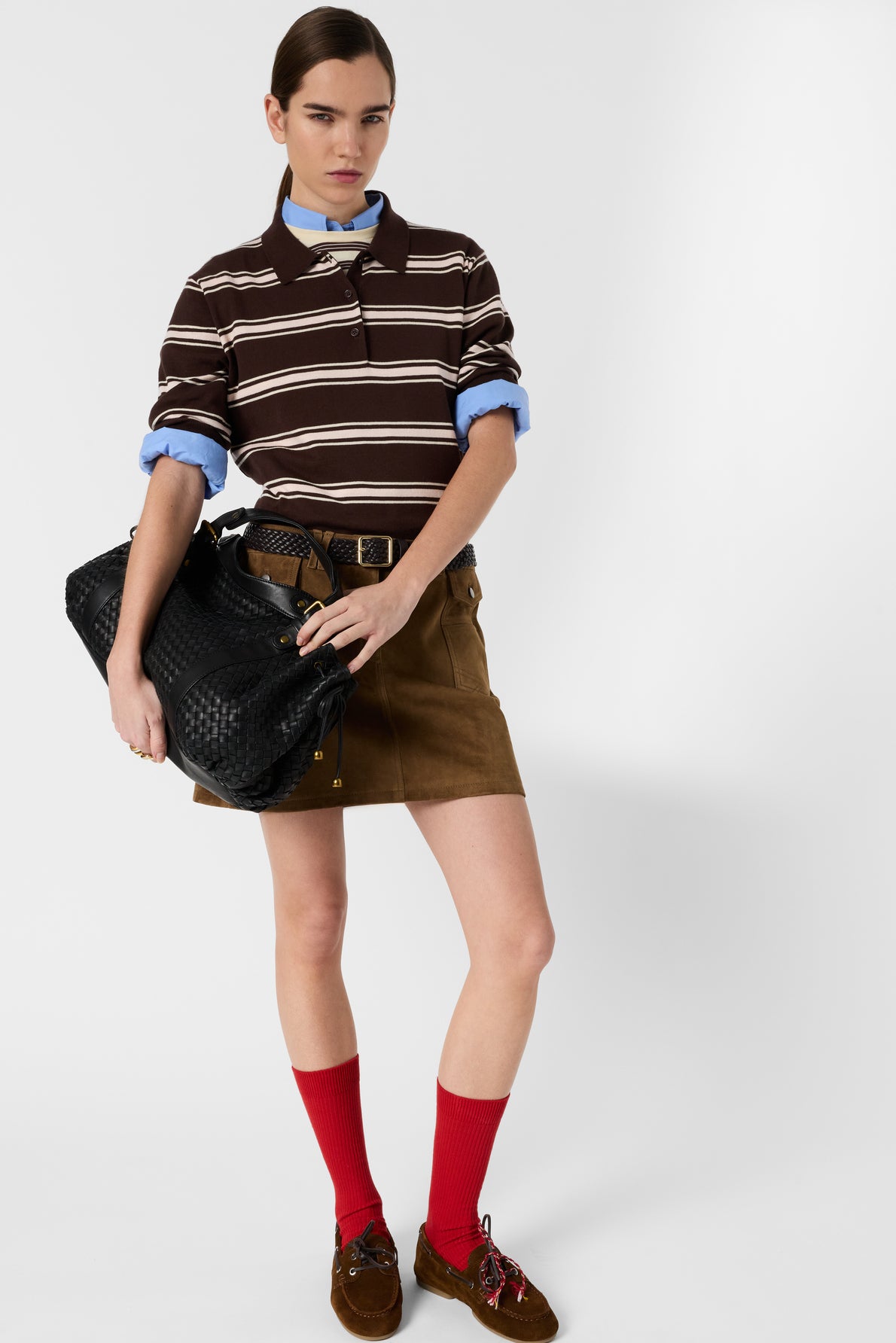 Woman in a brown striped rugby shirt reminiscent of a polo à rayures, blue shirt underneath, brown skirt, red socks, and brown shoes, holding a large black woven bag, standing against a plain white background.