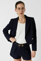 Woman wearing glasses, a navy blazer, navy shorts, and gold accessories poses with one hand in her pocket against a plain white background. She completes her look with a top en microfibre blanc, effortlessly blending comfort and sophistication.