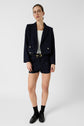 Person wearing a navy blazer, white top en microfibre blanc, navy shorts, black loafers with socks, glasses, and gold accessories stands against a plain white background.