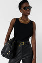 A person wearing black sunglasses, a chunky gold necklace, a sleeveless black top, high-waisted black leather pants with a gold-buckled belt, and holding a black leather bag.