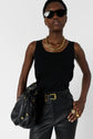 A person in a black tank top and leather pants poses with a large black bag, gold jewelry, and black sunglasses against a plain background.