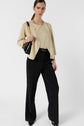 Woman standing in a beige blazer and top, black wide-leg trousers, and black heels, holding a black handbag with a gold chain, against a plain white background.