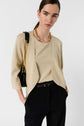 Woman with straight brown hair wearing a beige top, gold chain necklace, and black pants with a black belt. She has a black shoulder bag with a gold chain strap and stands against a plain light background.