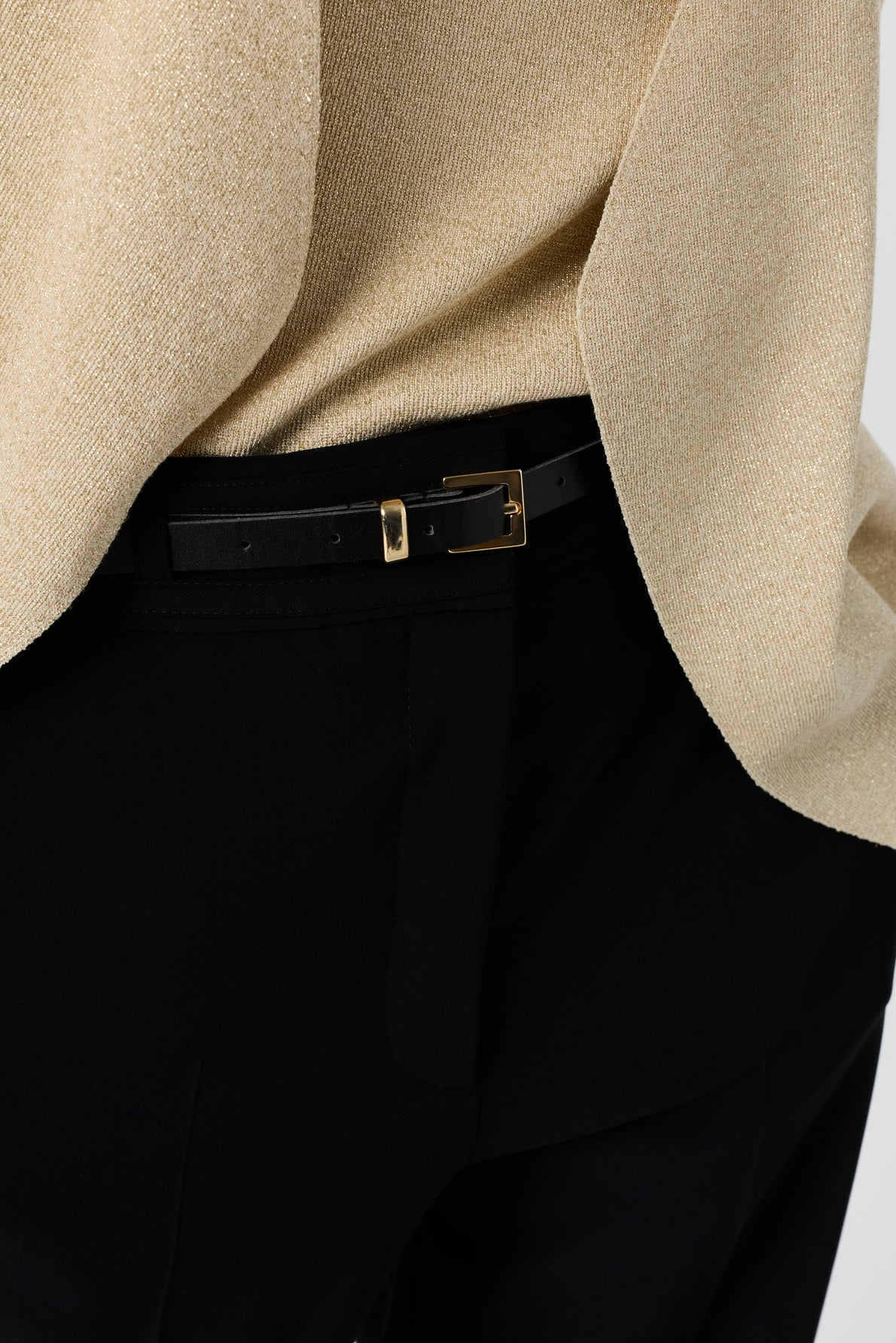 Close-up of a person wearing black pants with a black leather belt and a gold buckle, paired with a light beige, shimmery textured top.