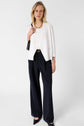 A woman stands against a plain background wearing a white blazer, black wide-leg trousers, black heels, and a gold necklace. She holds a black handbag and has a neutral expression.