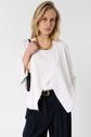 Woman with blonde hair wearing a white blazer, white top, black pants, and a gold chain necklace, holding a black shoulder bag. She stands against a plain light background, looking directly ahead.