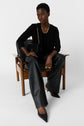 A person sits on a wooden chair, wearing a black blazer layered over a gilet femme chic, sleek black leather pants, pointed black heels, gold jewelry, and holding a black handbag with a gold chain. The background is plain white.