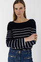 A woman with straight blonde hair wears a navy blue and white striped sweater and blue jeans. She stands with arms crossed, looking at the camera, against a plain white background.