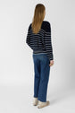 Person with long hair stands facing away, wearing a navy and white striped sweater, blue jeans, and white sneakers, against a plain light background.