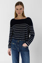 A person with straight, light brown hair wears a navy blue sweater with white horizontal stripes and blue jeans, standing with one hand in their pocket against a plain light background.