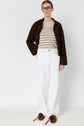 A person wearing glasses, a brown jacket, a pull à rayures, loose white pants, and brown loafers with tassels stands against a plain white background.