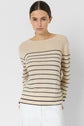 Woman wearing a beige pull à rayures with black horizontal stripes and white pants, standing against a plain light background, looking directly at the camera with a neutral expression.