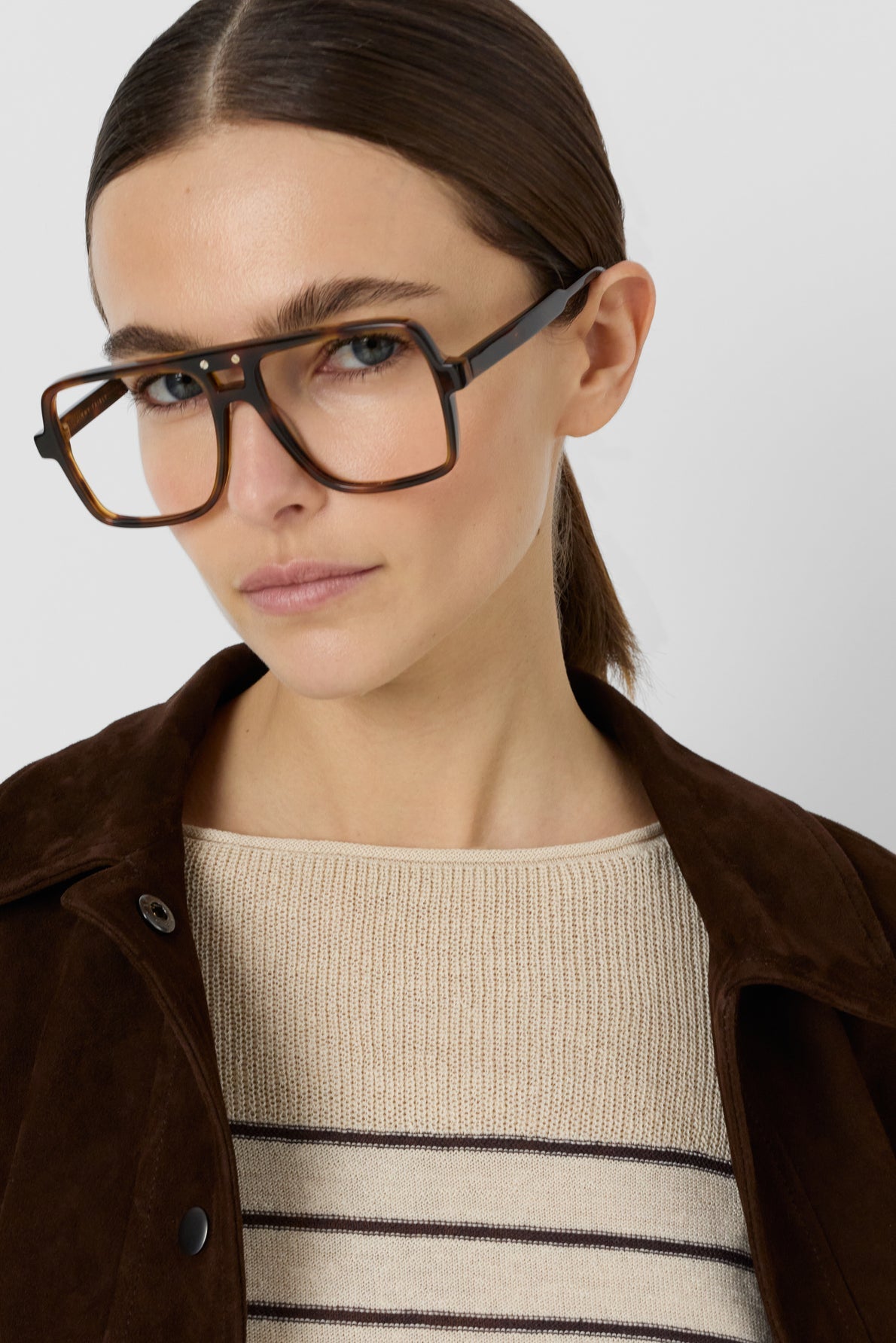 Woman with straight brown hair in a low ponytail wearing oversized square tortoiseshell glasses, a pull à rayures beige en lin mélangé, and a brown jacket, looking at the camera against a plain light background.