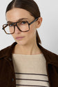Woman with straight brown hair in a low ponytail wearing oversized square tortoiseshell glasses, a pull à rayures beige en lin mélangé, and a brown jacket, looking at the camera against a plain light background.