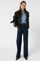 Woman standing against a white background, wearing glasses, a black leather jacket over a polo en lin à col polo ouvert et manches courtes, wide-leg dark jeans, and black pointed shoes, holding a black bag under her arm.