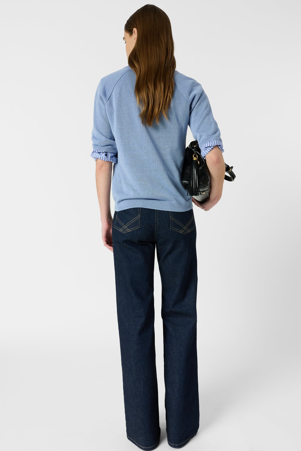 A woman with long brown hair stands facing away, wearing a light blue polo en lin with col polo ouvert and dark blue jeans, and holding a black handbag. The background is plain white.