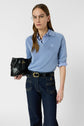 A woman with long brown hair wears glasses, a light blue polo en lin à col polo ouvert over a striped shirt, dark jeans with contrast stitching, and holds a black leather clutch. She stands against a plain white background.