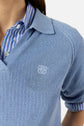 Person wearing a blue col polo ouvert knit shirt layered over a blue and white striped button-up shirt with manches courtes, featuring a small embroidered white logo on the chest.