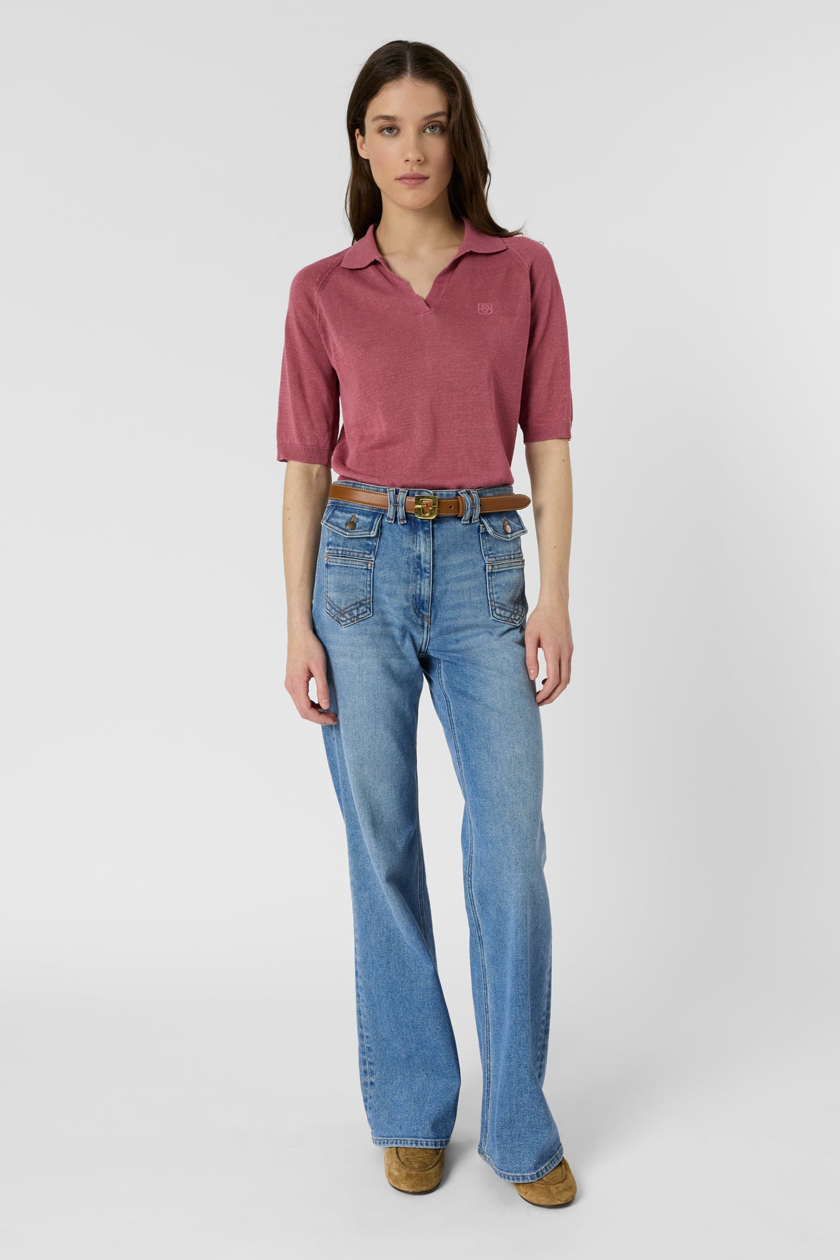 A woman stands facing forward, wearing a mauve polo lin femme with manches courtes, light blue wide-leg jeans with front pockets, a brown belt, and tan shoes against a plain white background.
