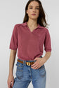 A woman stands facing forward, wearing a rose-colored polo manches courtes with a stylish collared neckline, tucked into high-waisted blue jeans with a brown belt. The background is plain and light-colored.