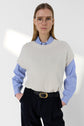 A person wearing glasses, a blue striped shirt, a white sweater vest sans manche with col rond, and black pants with a belt stands with hands in pockets against a plain light background.