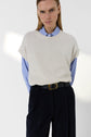 Person wearing a cream knit vest au col rond sans manche over a blue striped shirt, black trousers with a belt, and clear-framed glasses, standing against a plain white background—a chic layering piece reminiscent of the classic pull cachemire femme.