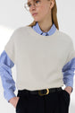 Person wearing a blue-and-white striped shirt under a cream knit vest sans manche, with black pants and a gold-buckled belt, looking to the side. They have glasses and straight, light brown hair, giving an effortlessly chic vibe reminiscent of a pull cachemire femme with a classic col rond.