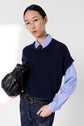 Person with long dark hair, wearing glasses, a blue striped shirt, and a pull cachemire femme bleu marine sans manche layered as a sweater vest, pairs the look with dark jeans and a black handbag while looking at the camera against a plain light background.