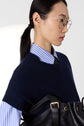 Woman in glasses wearing a bleu marine striped shirt, navy pull cachemire femme sans manche, and black tie, carrying a black leather bag with gold hardware, looking to the right.