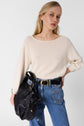 Woman with long blonde hair wears a cream sweater and high-waisted blue jeans with a black belt. She carries a large black handbag on her shoulder and stands against a plain white background, looking confidently at the camera.