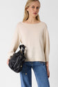 A woman with long blonde hair wears a cream sweater and blue jeans, carrying a black leather handbag over her shoulder, standing against a plain white background.