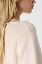 A close-up side view of a woman wearing a light beige knitted sweater, showing the shoulder and upper sleeve detail. Her long, blonde hair is partially visible.