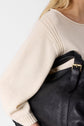 A person in a cream knit sweater holds a large black leather bag with a gold buckle strap under their arm. Only part of their upper body and hair are visible.