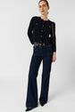 Woman standing against a plain background, wearing a navy gilet boutons dorés with gold buttons, high-waisted dark blue wide-leg jeans, black pointed shoes, and carrying a black shoulder bag with a gold chain strap.