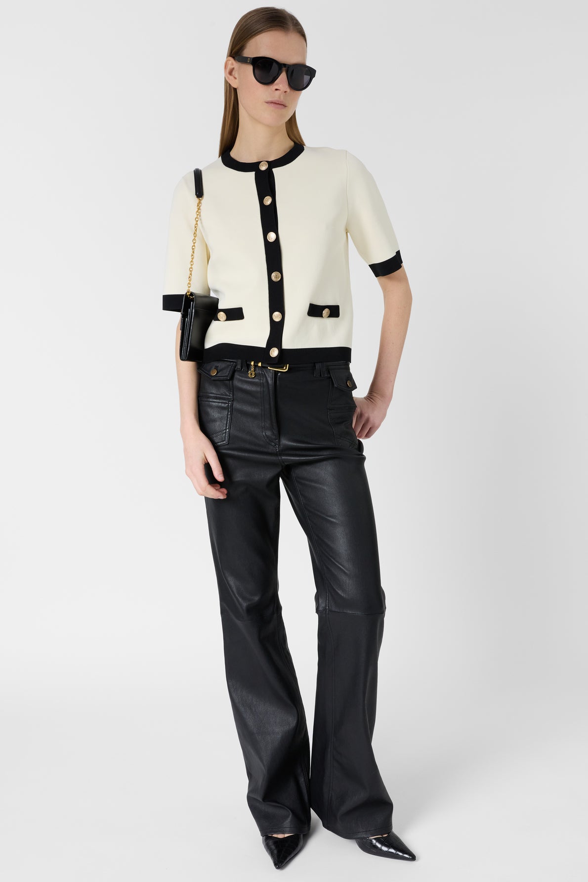 Woman wearing black sunglasses, a short-sleeve cream top with black trim and gold buttons layered over a gilet chic, black leather pants, and black pointed shoes, holding a black shoulder bag, standing against a plain white background.