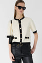A woman wearing sunglasses, a white short-sleeve top with black trim and gold buttons styled as a gilet chic, and black leather pants stands with one hand in her pocket and a small black purse across her body.